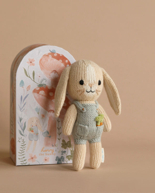 A Cuddle + Kind Tiny Henry The Bunny with long ears is wearing pale blue overalls and holding a small carrot plush. It stands in front of a decorative box featuring an illustration of the same bunny with mushrooms and floral designs, embodying sustainable fair trade practices.