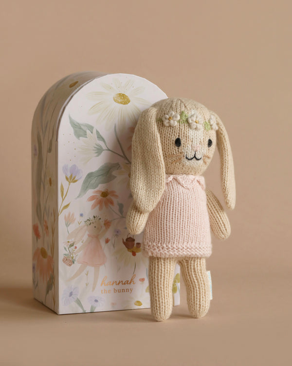 A Cuddle + Kind Tiny Hannah The Bunny with long ears, a pink dress, and flower decorations on its head is posed in front of a whimsical, floral-themed box. The handmade doll's box features illustrations of flowers and a girl, with the text "hannah the bunny" at the bottom.