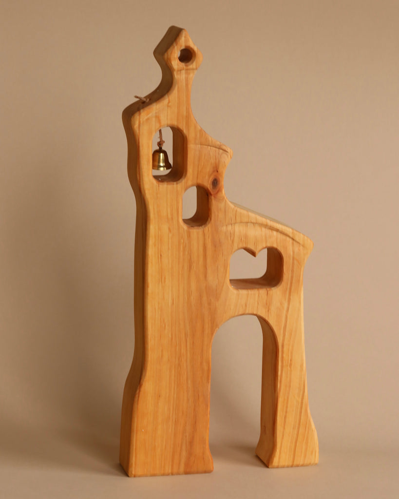 An Ostheimer Bell Tower resembling a stylized church or tower, with three hollow cutouts and a small bell hanging in the top cutout. This piece, made from sustainably sourced materials, has smooth edges and a natural wood finish, set against a plain beige background. Perfect for imaginative play.