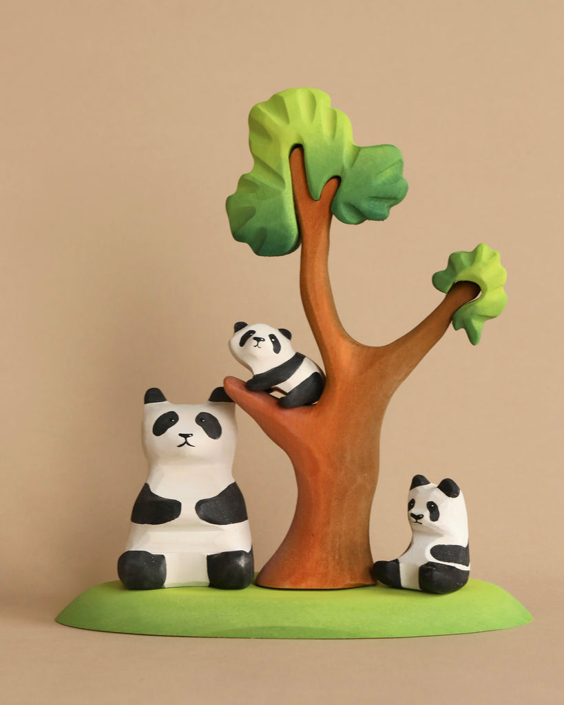 A Bumbu Panda Bears And Tree Set of three hand-carved panda bears and a tree on an eco-friendly wood base. One panda is sitting beside the tree, another is resting against it, and a smaller panda is positioned on a branch. The tree has a brown trunk and green foliage. Perfect for imaginative play scenes.