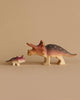 Two Bumbu Handmade Triceratops Dinosaur Set figurines, one larger and one smaller, resembling triceratops, crafted from sustainably sourced wood with brown and cream coloring. Purple-tipped frills and detailing highlight their features. Painted with non-toxic paint, they are set against a plain beige background facing each other.