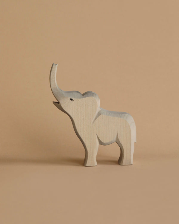 A light wooden toy elephant with its trunk raised stands against a beige background. Handcrafted using sustainable materials, this charming piece is reminiscent of the Ostheimer Small Elephant - Trumpeting, featuring simplicity and minimal details while casting a gentle shadow on the surface.