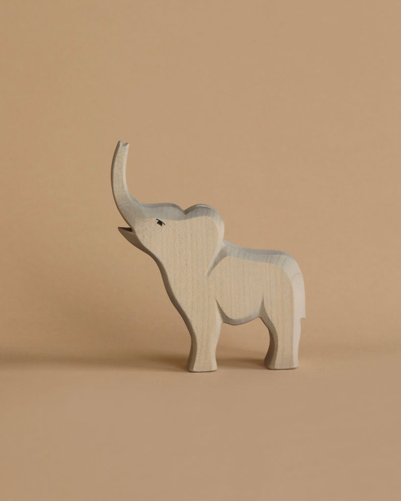 A light wooden toy elephant with its trunk raised stands against a beige background. Handcrafted using sustainable materials, this charming piece is reminiscent of the Ostheimer Small Elephant - Trumpeting, featuring simplicity and minimal details while casting a gentle shadow on the surface.