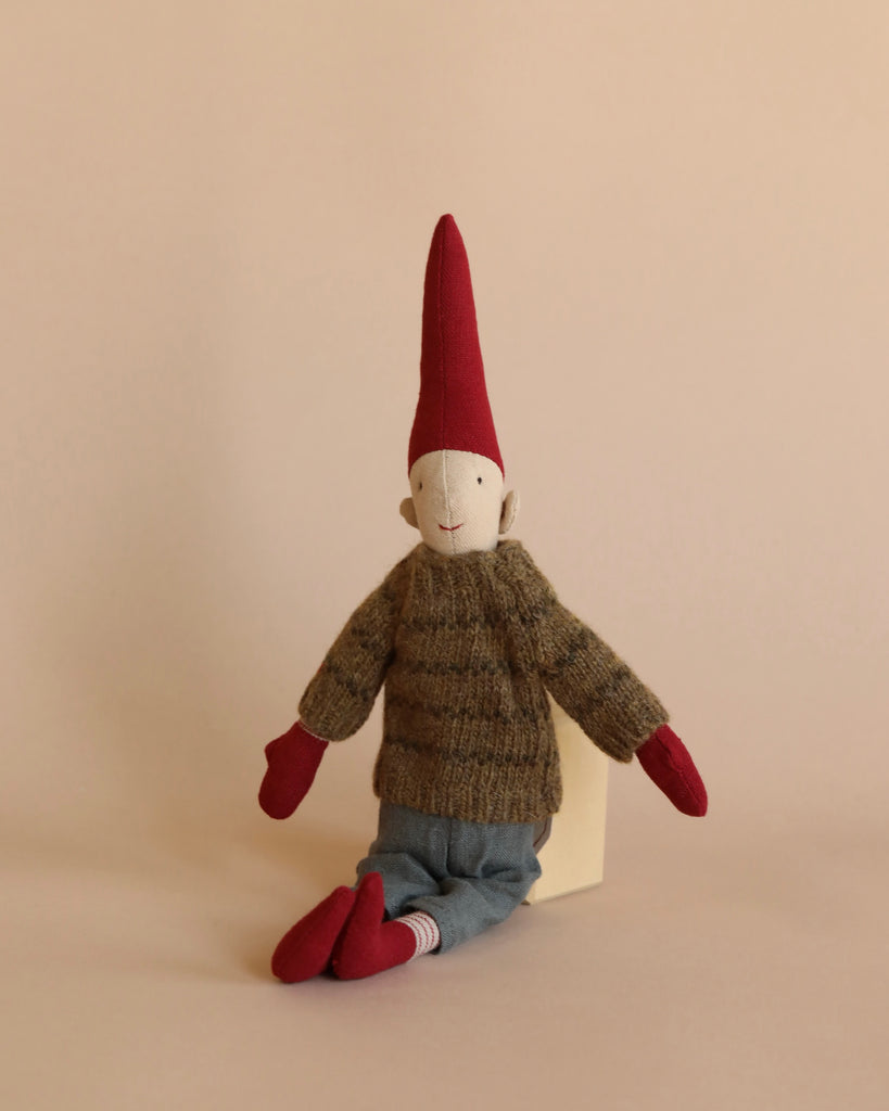 A Maileg Christmas Mini Pixy (Size 2), perfect as a Christmas decoration, sits against a beige background. The doll wears a pointy red hat, a thick brown knitted sweater made from exclusive materials, blue jeans, and red mittens and shoes. The neutral-faced mini pixie is propped upright, leaning slightly against the backdrop.