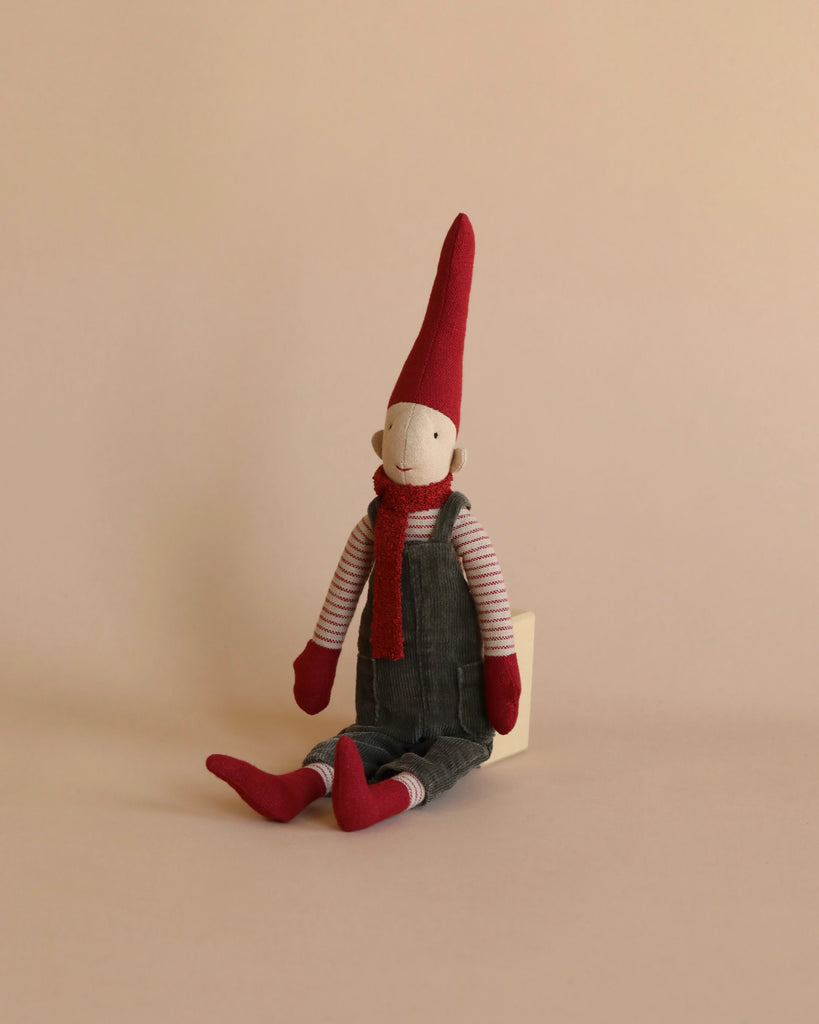 The Maileg Christmas Mini Pixy (Size 2) is a fabric doll adorned with a tall red hat, red scarf, red gloves, and red shoes. This charming Christmas decoration also features gray overalls and a striped shirt while sitting against a plain beige background.