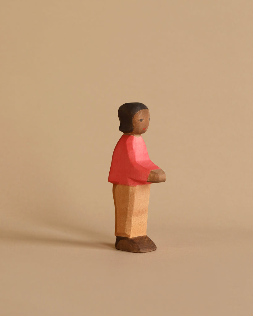 A small, handcrafted wooden figurine of a person stands against a beige background. The figure, dressed in a red sweater, tan pants, and brown shoes, has a simple, minimalist design with a smooth texture and neutral facial expression. Made using sustainably sourced materials, it evokes the charm of an Ostheimer Son, Red Sweater.