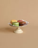 A small white pedestal stand holds four hand-painted Maileg Macarons, including yellow, green, brown, and pinkish-brown ones, placed on a delicate white doily. The scene could charm any of your Maileg Friends with its solid beige background.