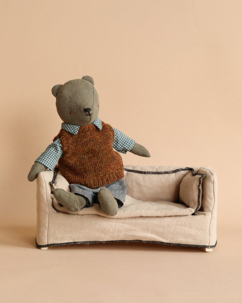 A handmade teddy bear, dressed in a brown sweater vest and blue checkered shirt, sits on a small beige fabric couch with black trim. The background is a plain beige, complementing the neutral tones of the scene. This charming "Maileg Extra Clothing: Shirt, Slipover and Pants for Teddy Dad" outfit can be purchased separately.