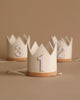 Three small Crown Party Hats, perfect for birthday celebrations and dress-up, are arranged in a row against a beige background. The front crown is white with a golden number 1, while the two crowns behind display numbers 2 and 3 in a similar style. Each party hat has a strap for wearing.