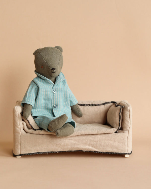 A stuffed bear, lovingly named Teddy Dad, is adorned in the Maileg Extra Clothing: Pyjama for Teddy Dad—a pair of striped blue pajamas—as it lounges on a small beige sofa against a neutral backdrop. This snug and charming outfit is available for purchase separately to enhance any cozy setting.