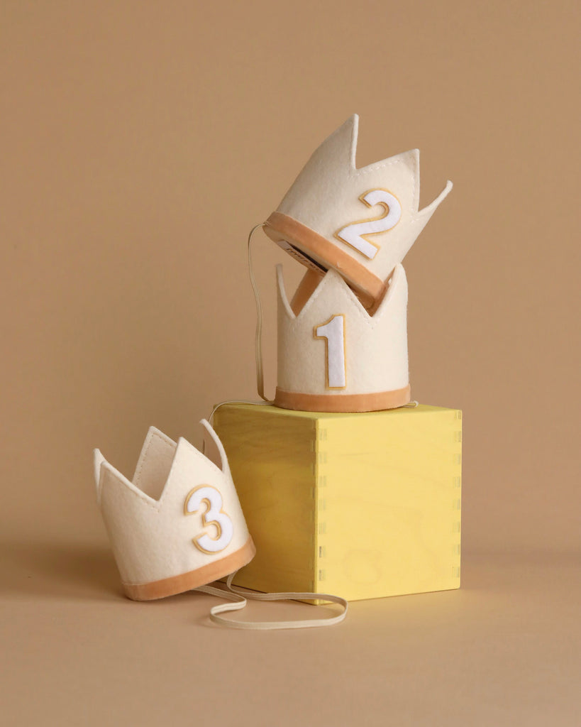 Three small, cream-colored felt Crown Party Hats, each with a gold number (1, 2, and 3), are arranged against a beige background. Perfect for birthday celebrations or dress-up fun, the first Crown Party Hat sits atop a yellow wooden block, the second is stacked on top of the first, and the third rests beside the block.