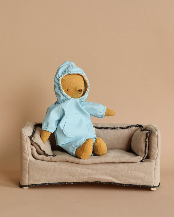 Teddy Junior, dressed in Maileg's extra clothing rainwear designed for him, sits comfortably on a small beige sofa against a plain background.