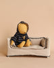 A small teddy bear, named Teddy Junior, is sporting the Maileg Extra Clothing: Duffle Coat for Teddy Junior and seated on a miniature beige sofa. The background and sofa are in neutral tones, creating a cozy and cute scene. Teddy Junior holds a tiny cup, adding to the adorable atmosphere.