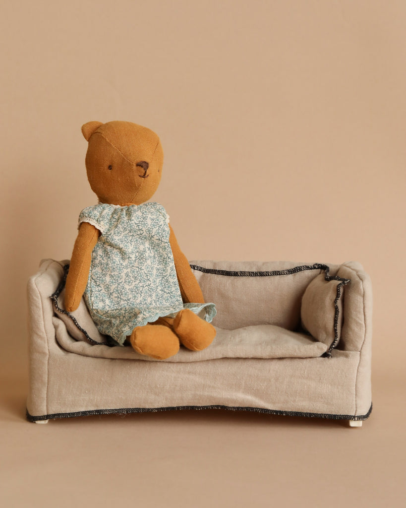 A brown Teddy Mum, wearing a Maileg Extra Clothing: Nightgown for Teddy Mum purchased separately, is seated on a small beige fabric sofa. The background is a plain, light brown color.