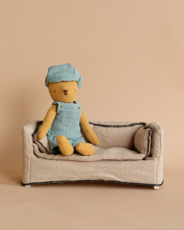 A small, stuffed teddy bear named Teddy Junior, adorned in a Maileg Extra Clothing overall and cap set, sits on a beige fabric sofa against a plain beige background. The teddy bear and the sofa are simple and softly textured, creating a cozy and minimalist scene.