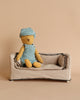 A small, stuffed teddy bear named Teddy Junior, adorned in a Maileg Extra Clothing overall and cap set, sits on a beige fabric sofa against a plain beige background. The teddy bear and the sofa are simple and softly textured, creating a cozy and minimalist scene.