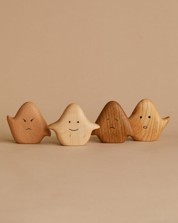 Four small Wooden Ghost Emotions, treated with natural linseed oil, are arranged in a row against a beige background. Each figurine displays one of the basic emotions: sad, happy, unhappy, and surprised.