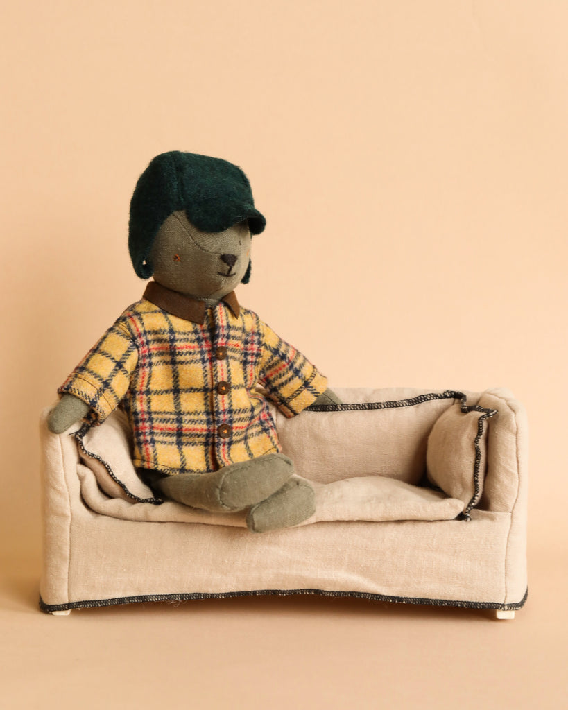 A plush bear, fondly called Teddy Dad, is dressed in a fashionable yellow plaid jacket and comfortably seated on a small beige sofa with dark trim. Adorned with a dark green hat and paws crossed in relaxation, this charming figure makes for a delightful addition to any collection. The outfit, known as Maileg Extra Clothing: Woodsman Jacket and Hat for Teddy Dad, and the sofa are available for purchase separately.