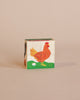 A Wooden Block Puzzle - Mini 4 Piece Domestic Animals painted with the image of a chicken standing on green grass, with an egg beside it, against a solid light pink background.