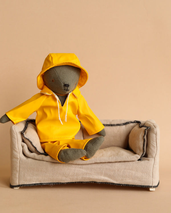 A plush Teddy Dad dressed in Maileg's charming rainwear outfit is seated on a small beige fabric couch, set against a plain tan background that highlights his readiness for rainy weather.