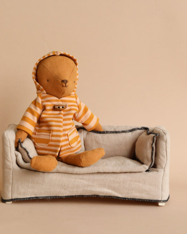 The Maileg Extra Clothing: Coat for Teddy Mum, a plush teddy bear in a snug orange hoodie with ocher stripes, sits on a small beige fabric couch against a neutral light brown backdrop.