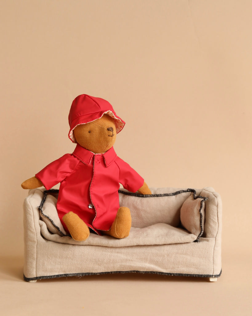 A teddy bear dressed in a red raincoat and hat, part of the charming "Teddy Mum" collection, sits on a small beige fabric couch. The background is a plain, light tan color. This adorable listing perfectly highlights the Maileg Extra Clothing: Rain Coat for Teddy Mum and its cozy outfit.