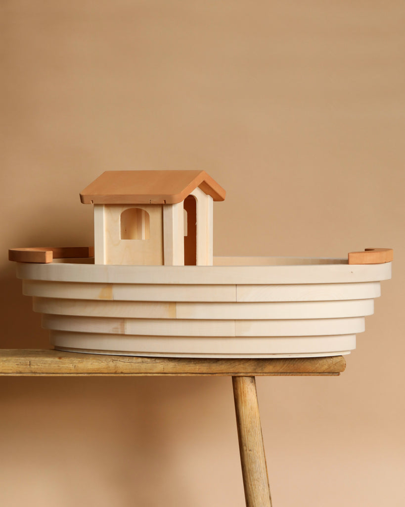 The Noah's Ark with Animals by Holztiger is a handmade wooden toy boat featuring a minimalist design and cabin. It rests elegantly on a wooden surface against a beige background, showcasing the beauty of solid wood craftsmanship.