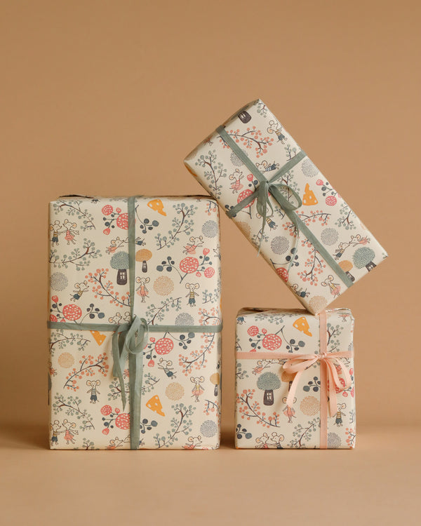 Three neatly wrapped gifts with colorful, whimsical wrapping paper featuring various patterns of plants, flowers, and mushrooms. The packages are tied with ribbon bows in different colors and arranged against a plain beige background, reminiscent of charming Maileg Mystery Box items that spark joy.