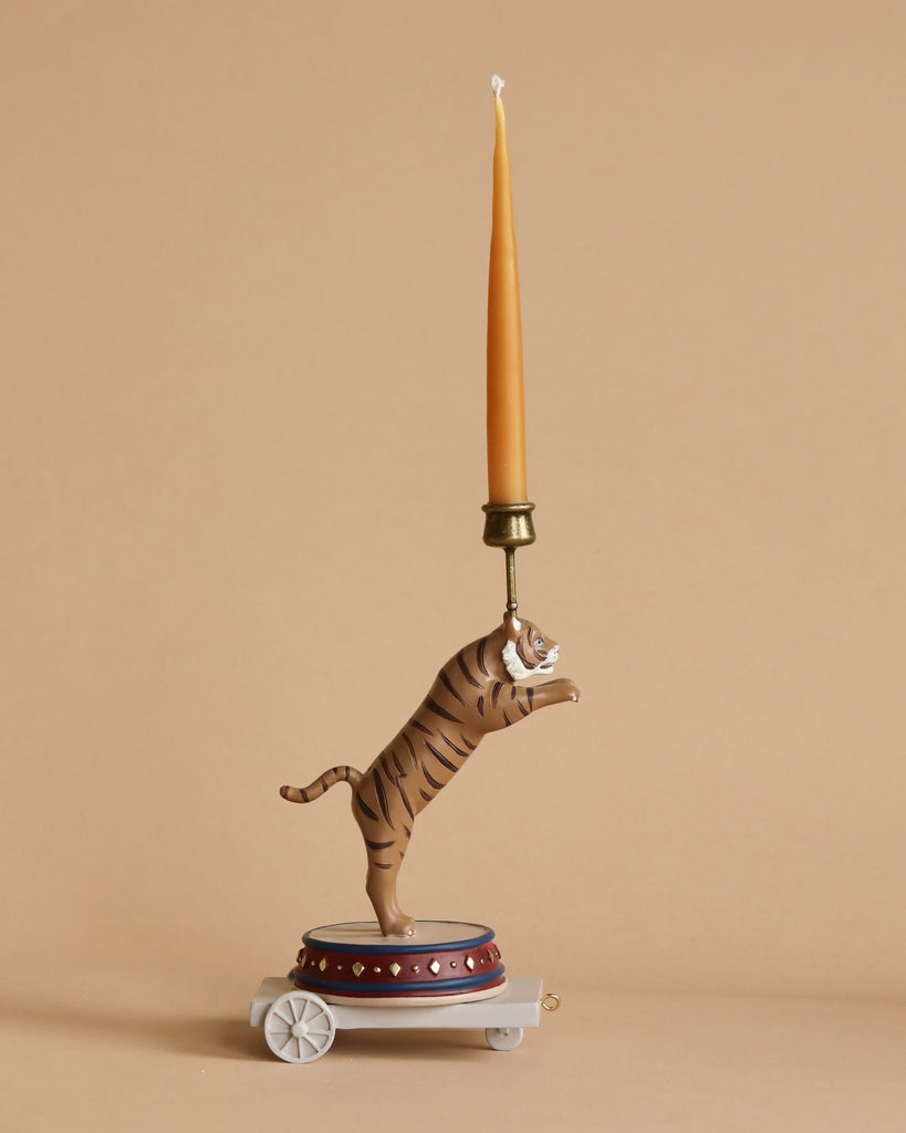 Introducing the Candle Holder - Tiger: a one-of-a-kind poly resin candle holder showcasing a standing tiger figurine on its hind legs, perfectly balanced on a blue and red circular base adorned with white wheels. The tiger is holding a tall, thin beeswax candlestick in its mouth, all set against a plain beige background.