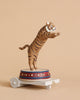 The Candle Holder - Tiger features a tiger figurine standing upright on its hind legs atop a round platform adorned with intricate gold and red details, reminiscent of an ornate poly resin candle holder. This platform is mounted on a small white wheeled cart, set against a plain beige background.