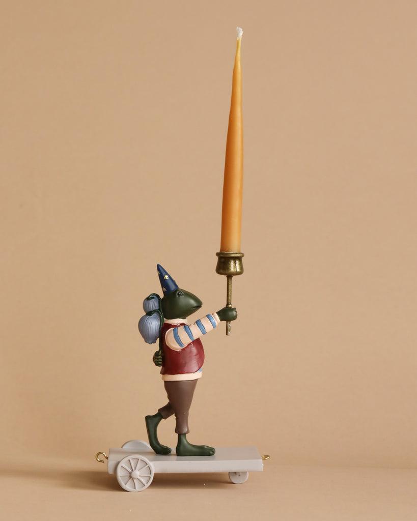 A figurine of a green, anthropomorphic creature with a long nose, wearing brown pants, a red vest, and a cone hat. Crafted from poly resin material, the "Candle Holder - Tiger" stands on a wheeled platform as part of a whimsical birthday train, holding a large tapered yellow candle upright in its right hand against a plain beige background.