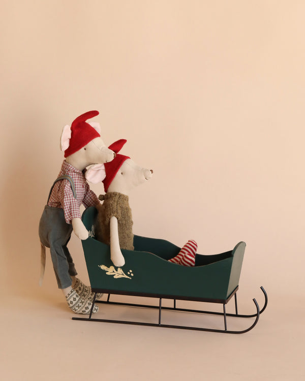 Two Maileg Medium Christmas Mice dressed in cozy winter clothing ride a green sled. One mouse, wearing a red hat and overalls, pushes from behind, while the other, in a red hat and green vest with red-striped socks, sits inside the medium-sized sled. The background is plain beige.