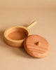 A Handmade Wooden Pretend Pan with a handle, resembling an heirloom-quality miniature saucepan, placed against a beige background. Made from fine hardwoods, the pan has a matching wooden lid with a knob on top, positioned slightly offset from the container—perfect for pretend play.