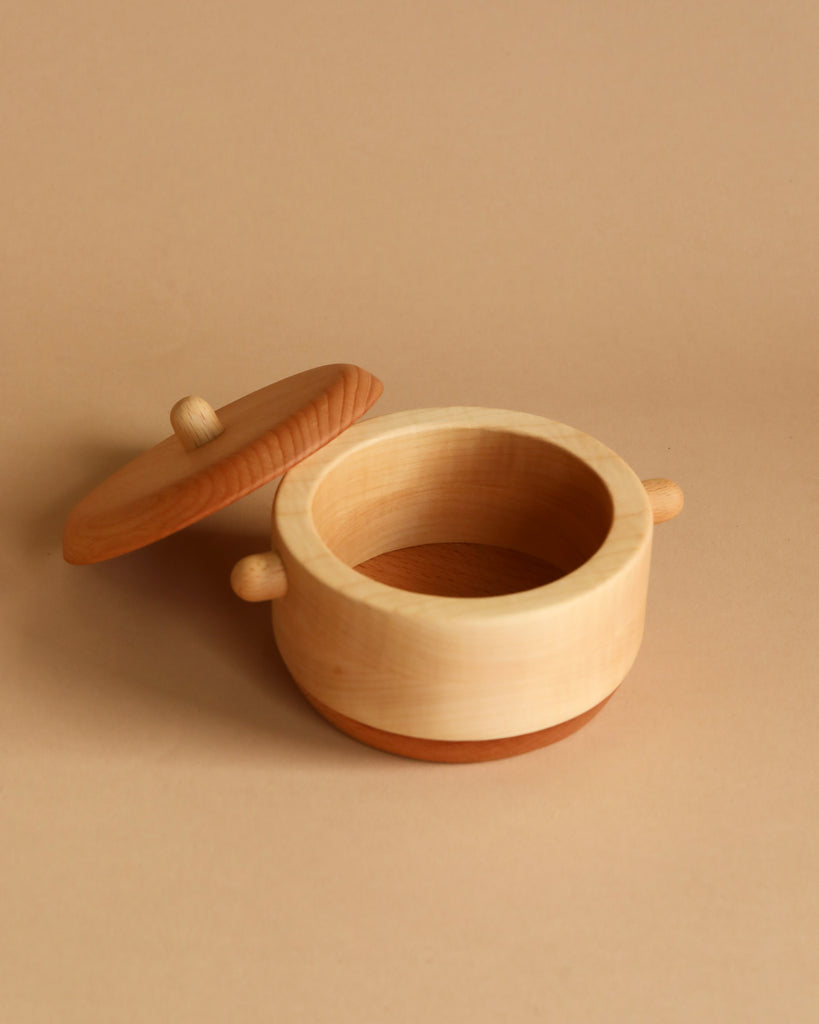 A round, light wooden container with a lid removed and placed to the side. The Handmade Wooden Pretend Pot has small handles on either side, and the interior is empty. Crafted from hardwoods like maple and beech, it sits against a uniform beige background.