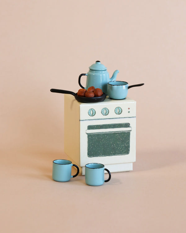 A miniature kitchen setup with a small white stove, topped with a blue kettle, a saucepan, and a skillet containing six small brown objects, likely vegetables or nuts. In front are three small blue mugs on a peach-colored background. This scene could be straight out of Maileg Madam Blue’s Favorites collection.