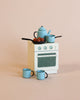 A miniature kitchen setup with a small white stove, topped with a blue kettle, a saucepan, and a skillet containing six small brown objects, likely vegetables or nuts. In front are three small blue mugs on a peach-colored background. This scene could be straight out of Maileg Madam Blue’s Favorites collection.