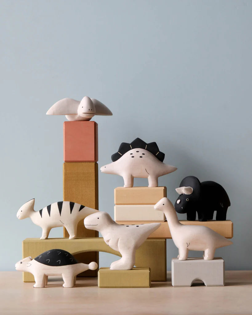 A collection of Handmade Tiny Wooden Dinosaurs - Ankylosaurus, including a seal, hippo, and others, neatly arranged on and around stacked pastel-colored blocks against a light blue background.