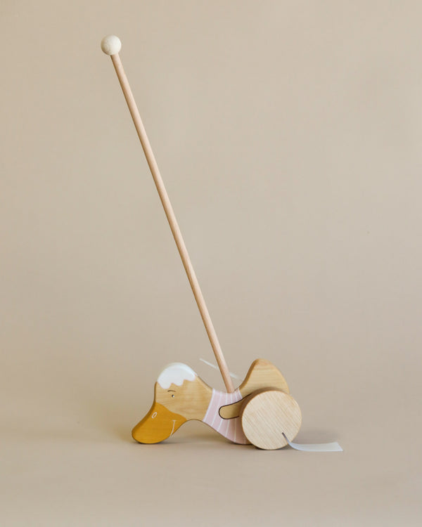 A delightful wooden push toy designed in the shape of a duck, expertly crafted from sustainably sourced birch wood. This toy showcases a natural wood finish with delicate white details on its head and wings, painted using natural milk paint. The extended handle and wheels ensure easy maneuverability, presented against a neutral beige backdrop. Officially named the "Handmade Wooden Duck Push Toy | Light Pink.