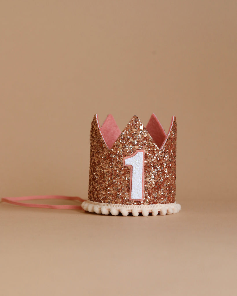 A small Rose Gold Glitter Crown with a pink interior sits on a beige background. The crown features a white number 1 on the front and has a pink ribbon attached to the sides. Perfect for birthday celebrations, the base of the crown is lined with small white pom-poms.