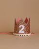 The Rose Gold Glitter Crown is a small, sparkling accessory with a pink interior and white pom-pom trim at the base. The crown showcases the number "2" in white and is set against a plain beige background. Pink ribbons on each side allow for easy tying, making it perfect for birthday celebrations.