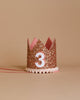 The Rose Gold Glitter Crown rests elegantly against a beige background, adorned with shimmering gold glitter and showcasing a white number "3" on the front. The crown's base is trimmed with delicate white pom-poms, while pink ribbon ties extend from the sides, making it perfect for birthday celebrations or party hats.