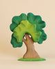 A whimsical sculpture of the Extra Large Wooden Tree crafted from linden wood with a vibrant green canopy and a brown trunk, set against a plain light beige background. The tree appears cartoonish with expressive features.