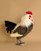 A realistic, life-sized Large Rooster Stuffed Animal stands upright against a plain beige background. This hand-sewn plush rooster boasts unique features like a red comb, white feathers around its head and neck, brown and white body feathers, a black tail, and gray feet.