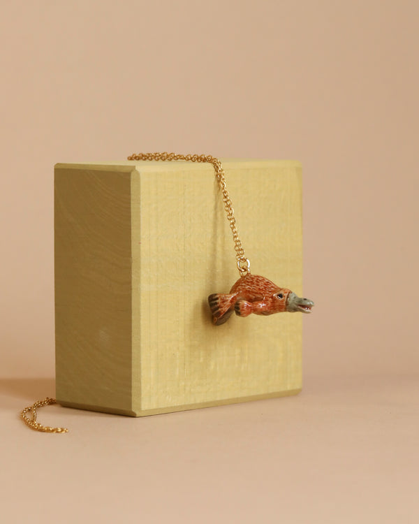 The Platypus Necklace showcases a refined, handcrafted porcelain pendant uniquely designed in the form of an orange fish with green accents. This exquisite piece is complemented by a 24k gold-plated steel chain, presented elegantly over a small, light-colored wooden box set against a neutral background.