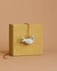 The Seal Necklace features a white porcelain pendant shaped like an adorable seal, suspended on a gold chain. This heirloom-quality piece is elegantly displayed over a small wooden block with a light yellow finish, set against a beige background.