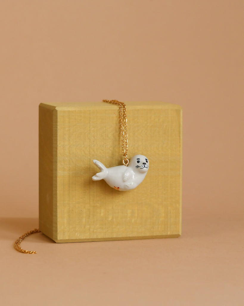 The Seal Necklace features a white porcelain pendant shaped like an adorable seal, suspended on a gold chain. This heirloom-quality piece is elegantly displayed over a small wooden block with a light yellow finish, set against a beige background.