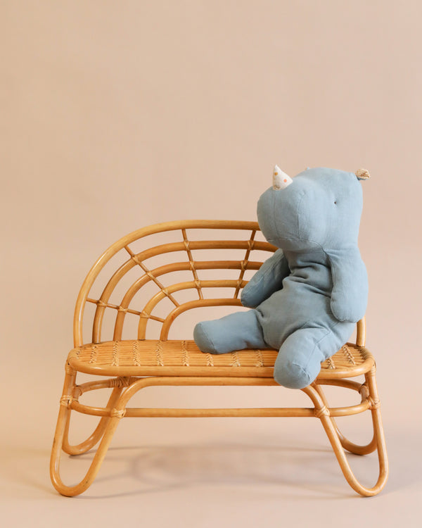 A soft, blue stuffed animal resembling a rhinoceros is seated on a small, curved Kid's Rattan Bench against a neutral background. The plush toy has a white horn and a simplistic design, adding a cute and cozy charm to the scene.