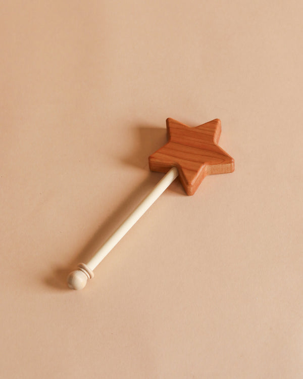 A Bannor Toys Wooden Star Wand with a star-shaped tip and a long handle, laying on a plain pastel beige background.