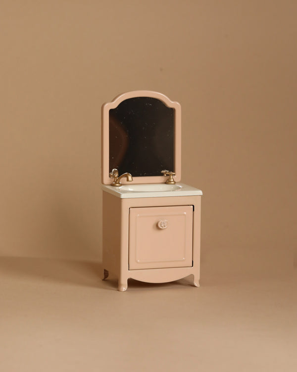 The Maileg Sink Dresser With Mirror, Mouse - Powder, featuring a beige finish, curved top mirror, gold-colored faucets, white sink, and small storage cabinet beneath, is perfect for a mouse house and positioned against a neutral background.