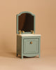 The Maileg Sink Dresser With Mirror, Mouse - Mint is pictured. This small, vintage-style dollhouse vanity in a soft green color features a single cabinet door with a gold knob. The top surface includes a white sink with golden faucet and handles. The background is plain beige.